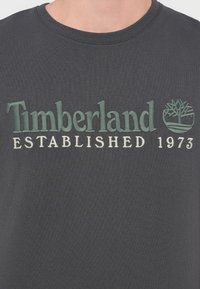 Σκούρο γκρι βαμβακερό μπλουζάκι με κεντητό κείμενο «Timberland ESTABLISHED 1973» σε πράσινο και λευκό, μαζί με λογότυπο δέντρου.