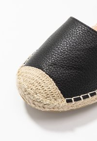 Espadrille de cuero negro con puntera de ganchillo beige y suela de yute, mostrando material texturizado y detalles de costura sobre fondo blanco.