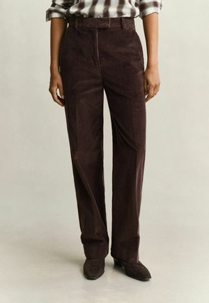 Personne portant un pantalon en velours côtelé marron foncé taille haute avec passants de ceinture et des bottines en daim marron, debout sur un sol clair et uni.