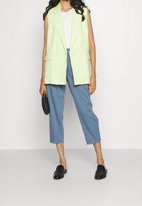 Jasnozielona bezrękawnikowa marynarka z dwoma przednimi kieszeniami, zestawiona z luźnymi niebieskimi spodniami o długości 3/4, czarnymi wsuwanymi butami i małą czarną torebką.