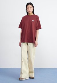 Une femme se tient devant un fond blanc uni, portant un t-shirt couleur rouille et un pantalon beige à jambes larges, associé à des baskets.