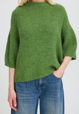 Personne portant un pull tricoté vert à manches trois quarts et un jean bleu, une main dans la poche, sur un fond uni.