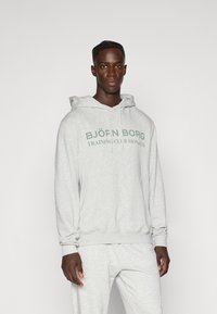 Γκρι φούτερ με καγκουρό τσέπη μπροστά και ρυθμιζόμενο κορδόνι. Διαθέτει την ένδειξη "BJÖRN BORG TRAINING CLUB MONACO" εκτυπωμένη σε σκούρο πράσινο.