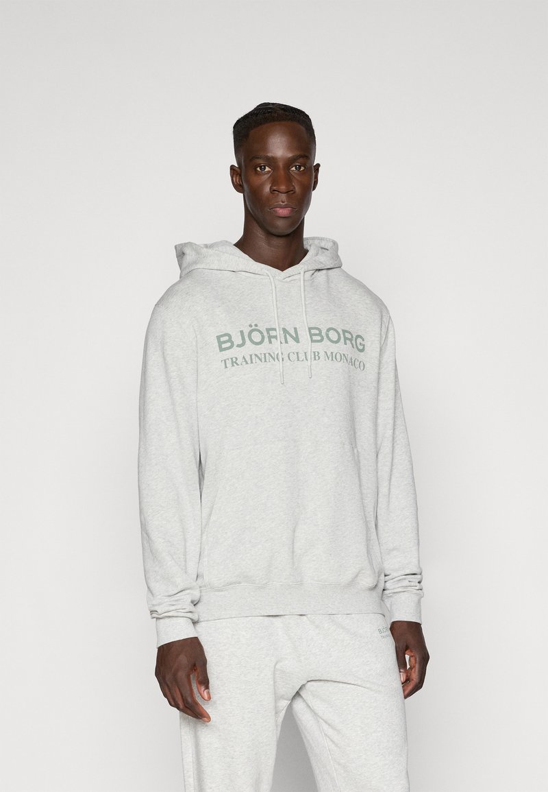 Γκρι φούτερ με καγκουρό τσέπη μπροστά και ρυθμιζόμενο κορδόνι. Διαθέτει την ένδειξη "BJÖRN BORG TRAINING CLUB MONACO" εκτυπωμένη σε σκούρο πράσινο.