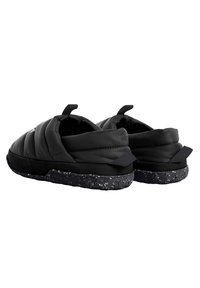 Musta pörröiset tossut, joissa on teksturoitu päällinen, rosoinen musta ja valkoinen pohja, vetotapit takana ja ainutlaatuinen siluetti.