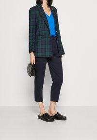 Groene en marineblauw geruite blazer, blauwe V-hals top, zwarte cropped broek en zwarte instapschoenen. Houdt een gestructureerde zwarte handtas vast.