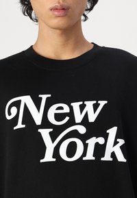 Čierna mikina vyrobená z bavlny s veľkým bielym nápisom "New York." Okrúhly výstrih a voľný strih, s krátkymi viditeľnými rukávmi.