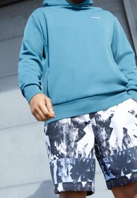 Persona con una sudadera teal de Calvin Klein y pantalones cortos de patrón blanco y negro, de pie al aire libre frente a una pared gris.