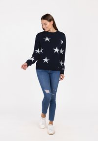 Navy Pullover mit weißem Sternenmuster, Rundhalsausschnitt und langen Ärmeln, kombiniert mit blauen, zerrissenen Jeans und weißen Sneakers.