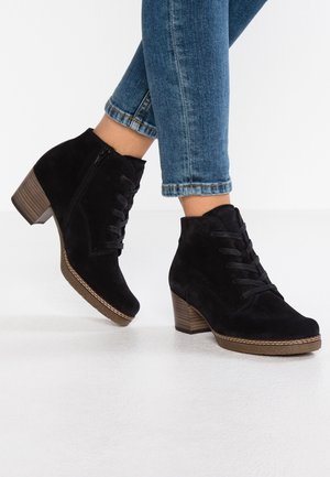 Bottines en daim noir à talons carrés et fermeture éclair latérale, portées avec un jean bleu retroussé sur un sol blanc.