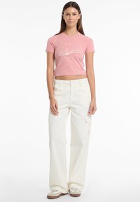 Žalioji rožinė marškinė su balta grafika, derinama su laisvo kirpimo baltais kargo džinsais, turinčiais šonines kišenes ir mygtukų užsegimą.