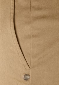 Tissu de pantalon en coton beige avec une texture en rib tout en diagonale, poche latérale et un bouton métallique circulaire gravé "MAISON SCOTER".