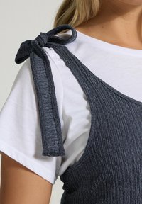 Mörkgrå texturerad linne med ärmlös design, med en knyt-detalj på axeln, lager-på-lager över en vit t-shirt.