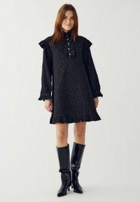 Czarna sukienka z długim rękawem, z falbanami i wysokim kołnierzem. Posiada zapięcie na guziki oraz teksturowany kwiatowy wzór. W zestawie z błyszczącymi butami sięgającymi za kolano.