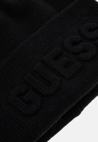 Cappello in maglia nera con il logo "GUESS" testurizzato in evidenza. Materiale morbido, forma arrotondata e dettaglio con risvolto.