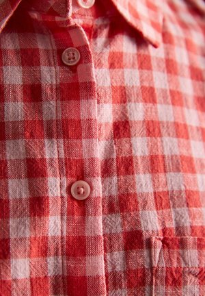 Sarkanas un baltas rūtainas pogājamas krekls ar apkakli un redzamām plastmasas pogām, ataino auduma tekstūru un raksta detaļas.