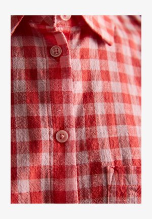 Czerwona i biała koszula w kratę na guziki, z kołnierzykiem i widocznymi plastikowymi guzikami, ukazująca teksturę tkaniny i detale wzoru.
