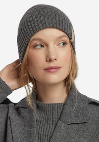 Graue gerippte Strickmütze mit umgeschlagenem Bund und einem kleinen cremefarbenen Etikett. Getragen mit einem grauen Pullover und Mantel, die eine glatte Textur zeigen.