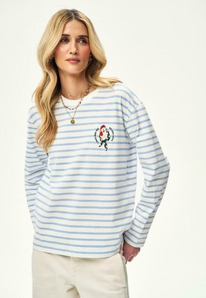 Blondynka w jasnoniebiesko-białej koszuli w paski z długim rękawem, z haftem koguta na piersi, w beżowych spodniach, stojąca na białym tle.