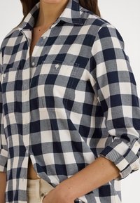 Žena oblečená v tmavomodrej a bielej károvanej košeli s gombíkmi, rukávy vyhrnuté, s náprsnými vreckami, v kombinácii so svetlými béžovými nohavicami.