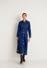 Kvinne iført knappet mørkeblå denimjakke og matchende midiskjørt, holder svart håndveske, går mot en enkel hvit bakgrunn.