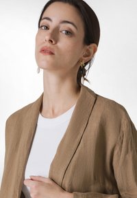 Ruskea pellavapusero, jossa on rento leikkaus valkoisen puuvillat-paidan päällä. Kultaiset ja mustat roikkuvat korvakorut, joissa on helmiakseli. Neutraali tausta.