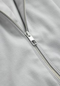 Close-up of a silver metal zipper partially undone on a light gray fabric surface, showing detailed zipper teeth and pull tab.