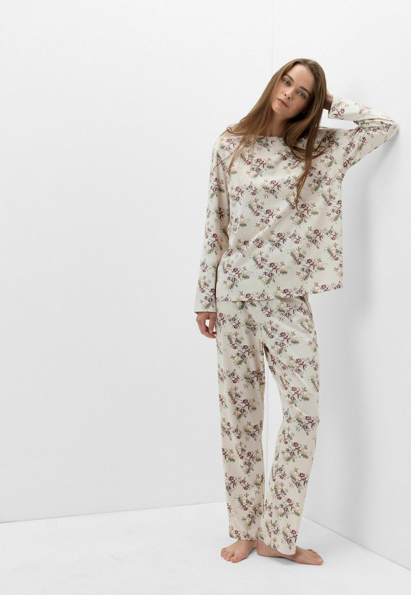 Young woman with long hair wearing cream floral long-sleeve top and matching pants, standing barefoot against a white wall.