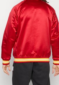 Giacca bomber in raso rosso con colletto a coste, polsini elastici e orlo con strisce gialle e bianche. Texture liscia, design casual.