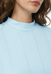 Helles blaues Oberteil mit Stehkragen aus geripptem Stoff, das dezente vertikale Linien und eine figurbetonte Silhouette aufweist. Weiche Textur und minimalistisches Design.