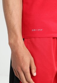 Czerwona koszulka sportowa DRI-FIT z fakturowanym materiałem. Zbliżenie pokazuje detale szwów oraz ramię, z widocznymi poniżej czarnymi szortami o fakturze.