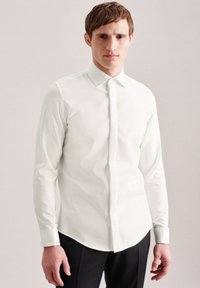 Camicia bianca a maniche lunghe con colletto a punta, slim fit, tessuto liscio e leggero lucido. Pantaloni scuri abbinati alla camicia.