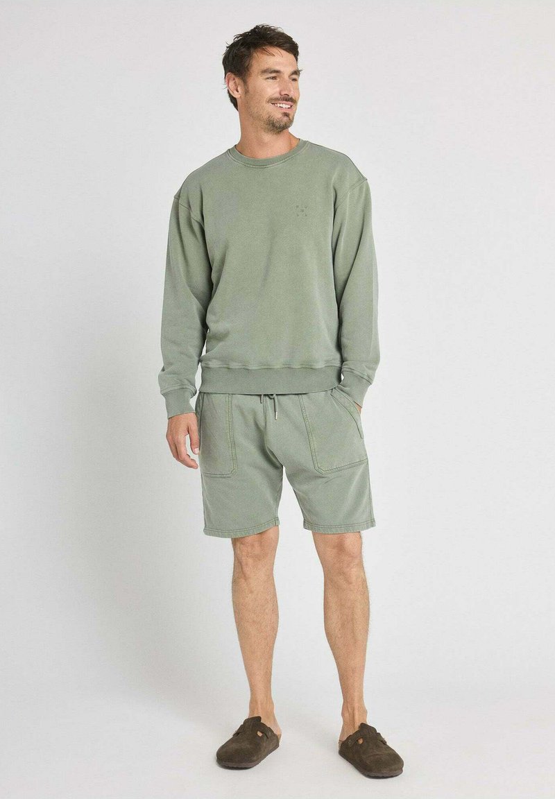 Homme debout, portant un sweat vert sauge et un short assorti avec des poches, ainsi que des chaussures marron foncé sans lacets, souriant et regardant vers la droite.