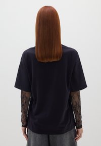 Schwarzes, übergroßes T-Shirt mit kurzen Ärmeln, überlagert mit durchsichtigen schwarzen Spitze-Ärmeln. Es bietet einen entspannten Schnitt und eine glatte Textur.