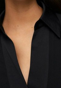 Schwarzes Shirt mit spitzem Kragen und tiefem V-Ausschnitt, mit glatter Textur und halb transparenter Optik. Zwei sichtbare kleine Punkte auf der Haut.