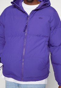 Fialová péřová bunda se zipem vpředu, kapucí a bočními kapsami; má hladký, lesklý povrch a nenápadné označení na hrudi.