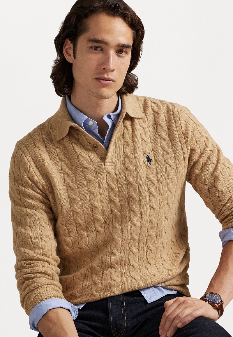 Pull en tricot beige avec col, orné d'un petit logo marine. Porté par-dessus une chemise à boutons bleu clair, avec une montre au poignet.