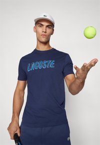 Mornarica majica s kratkimi rokavi z napisanimi "LACOSTE" v svetlo modri barvi. Model nosi belo kapo in drži teniško lopar; teniška žogica lebdi nad njim.