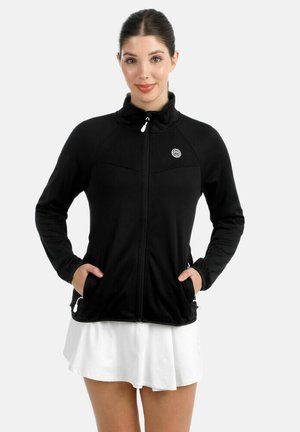 Vrouw met een zwart jack met ritssluiting en handen in de zakken, en een witte rok, staand tegen een effen lichte achtergrond.