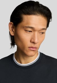 T-shirt girocollo in cotone nero con colletto a coste a strisce bianche e grigie. Il modello ha capelli scuri e lisci e un'espressione neutra.