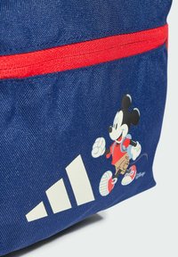 Mochila de tejido azul marino con un zipper rojo, que presenta un gráfico de Mickey Mouse y acentos del logo blanco de Adidas. Superficie texturizada.