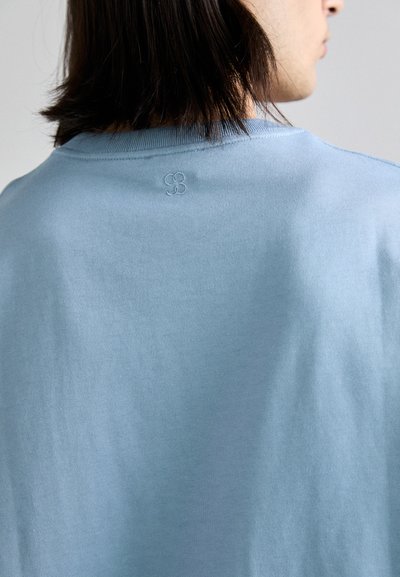 Camiseta de algodón azul claro con cuello redondo y un sutil logo bordado en la parte posterior. Textura suave y mangas cortas.