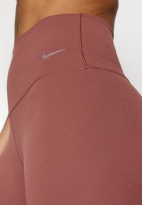 Bordó, magas derekú leggings sima textúrával, egy kis szürke Nike logóval a derékpánton és kiemelkedő varrás részletezéssel.