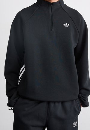 Person iført sort Adidas træningsdragt med hvidt logo, quarter zip sweatshirt og bukser med tre hvide striber på siden.