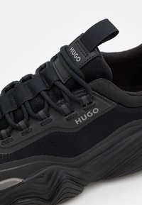 Sneaker nera con tomaia in rete, sovrapposizioni in tessuto e suola in gomma spessa. Il marchio "Hugo" è impresso sulla linguetta e sul lato.
