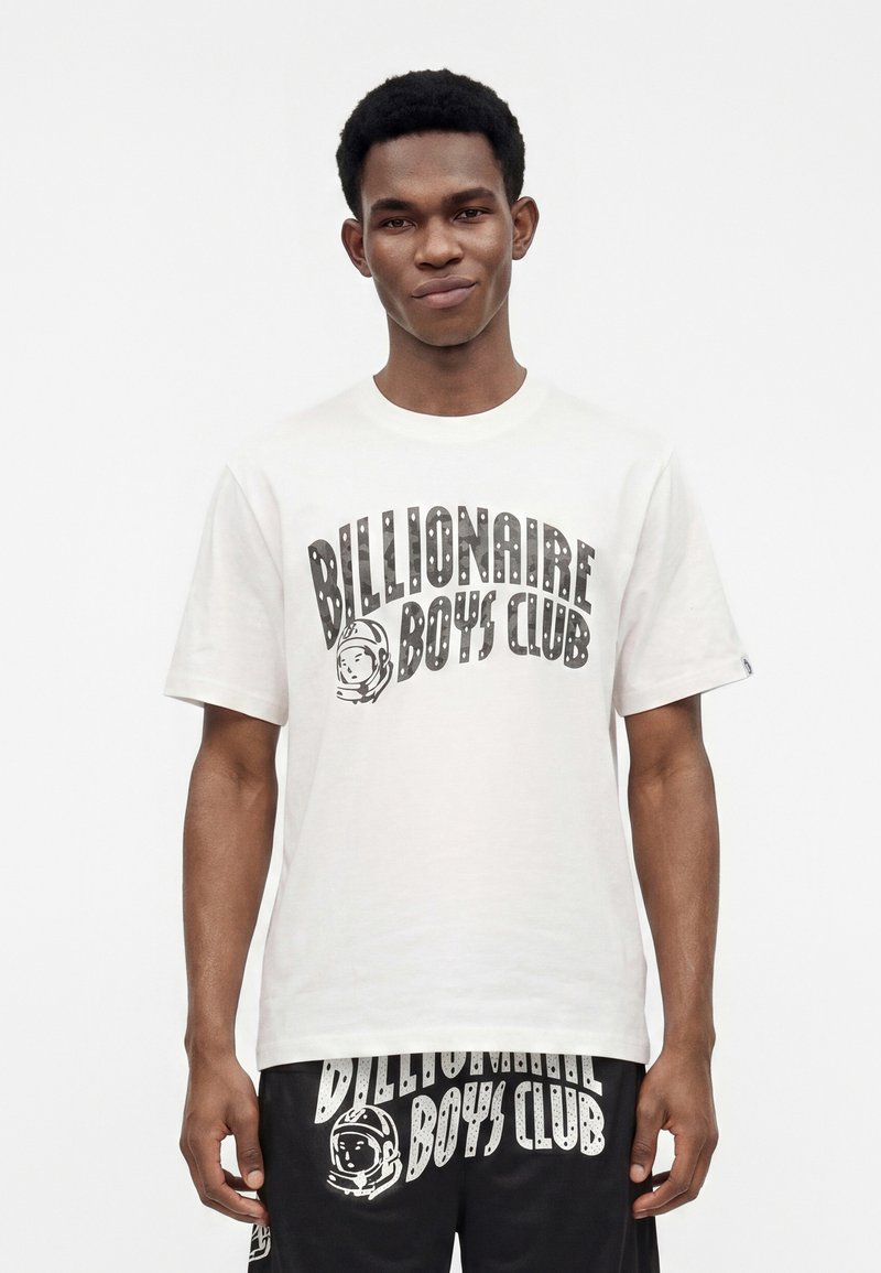 Νέος άνδρας που φοράει λευκό μπλουζάκι Billionaire Boys Club και μαύρο σορτς με αντίστοιχα σχέδια λογότυπου, στέκεται μπροστά σε μια απλή λευκή φόντο.