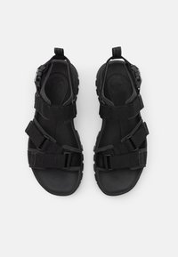 Sorte sandaler med justerbare stoffstropper, gummisåle og teksturert innersåle. Har en sikker bakstropp for stabilitet.