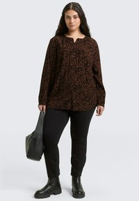 Woman wearing a brown and black animal print blouse, black pants, black boots, and holding a black handbag, standing against a light background.