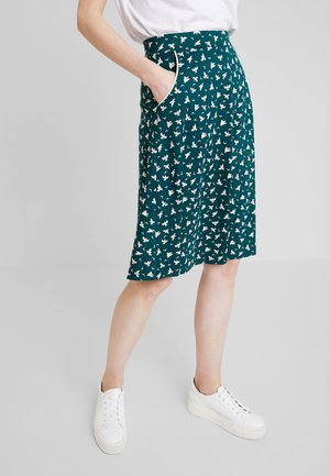 Vrouw draagt een teal-kleurige knielange rok met witte vogelpatronen, witte sneakers en een ingestopt wit topje, staand met één hand in de zak.