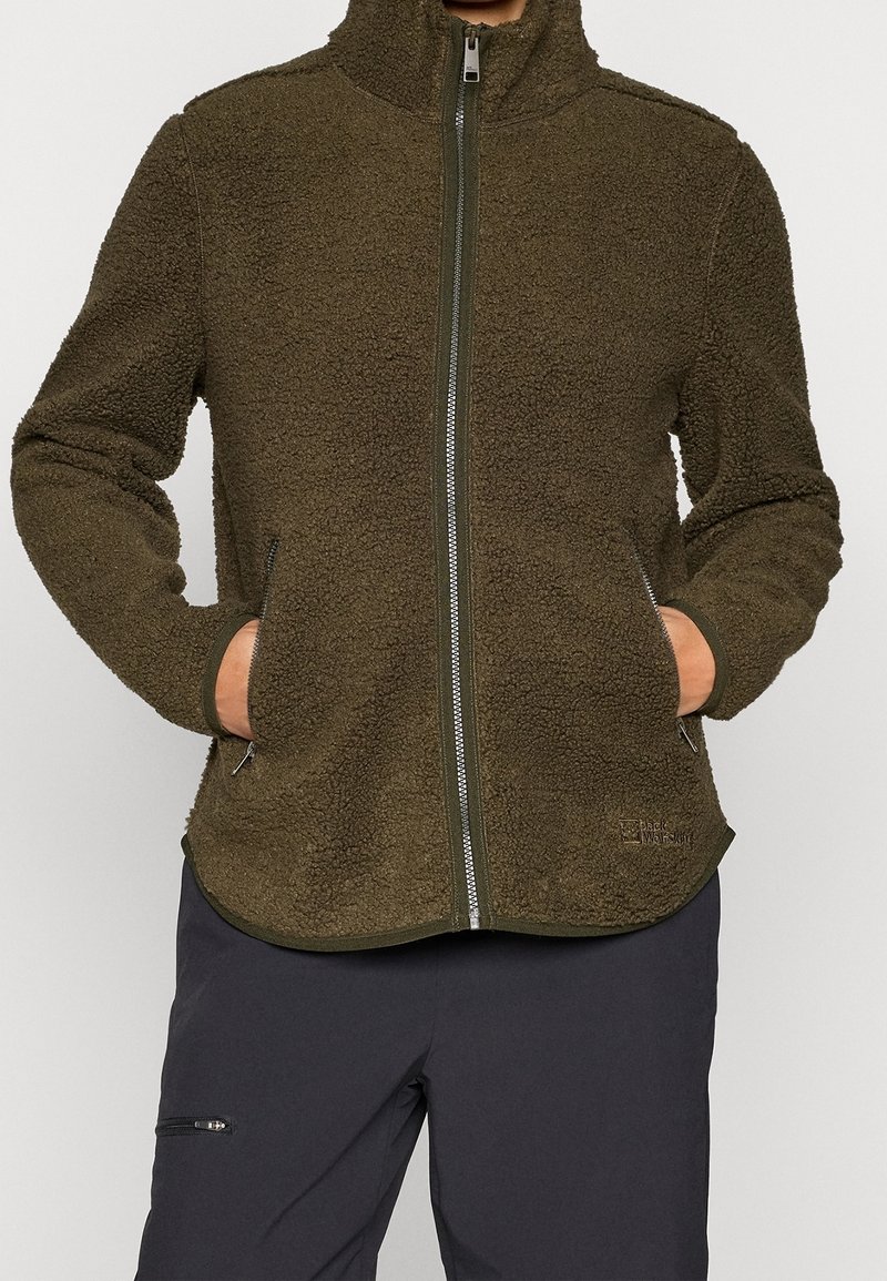 Personne portant une veste polaire zippée vert olive avec les mains dans les poches latérales et un pantalon foncé avec une poche zippée à la cuisse.